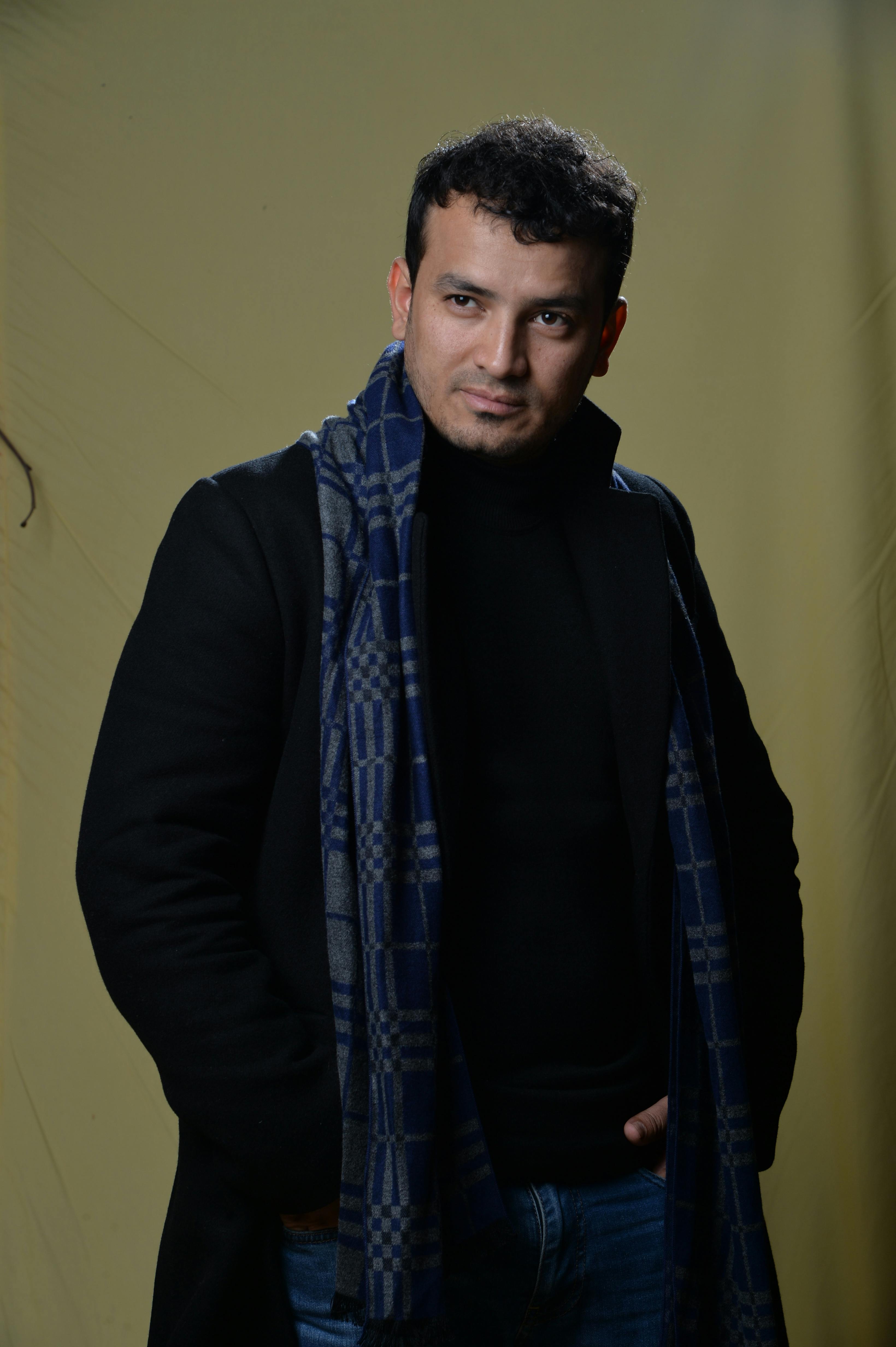 Confident man in black blazer and scarf smiling warmly against a neutral backdrop.