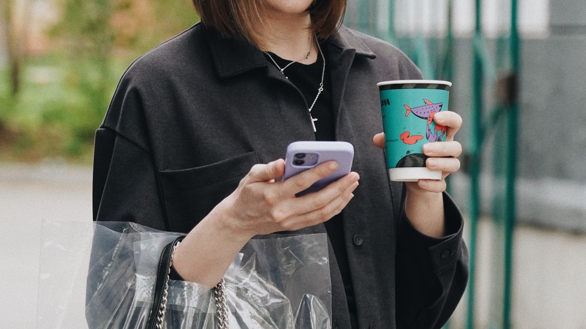 A young woman checking her phone on a city street, holding a coffee and a shopping bag