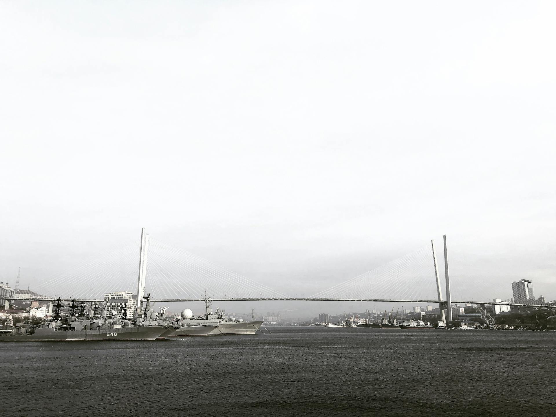 Black and white photo of Vladivostok harbor featuring docked ships and a suspension bridge.