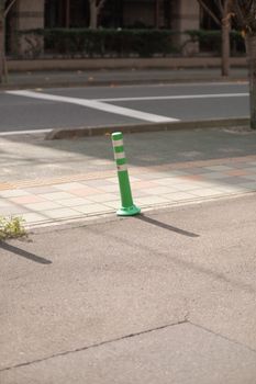 A green bollard stands alone on a sunny city sidewalk, casting a shadow.
