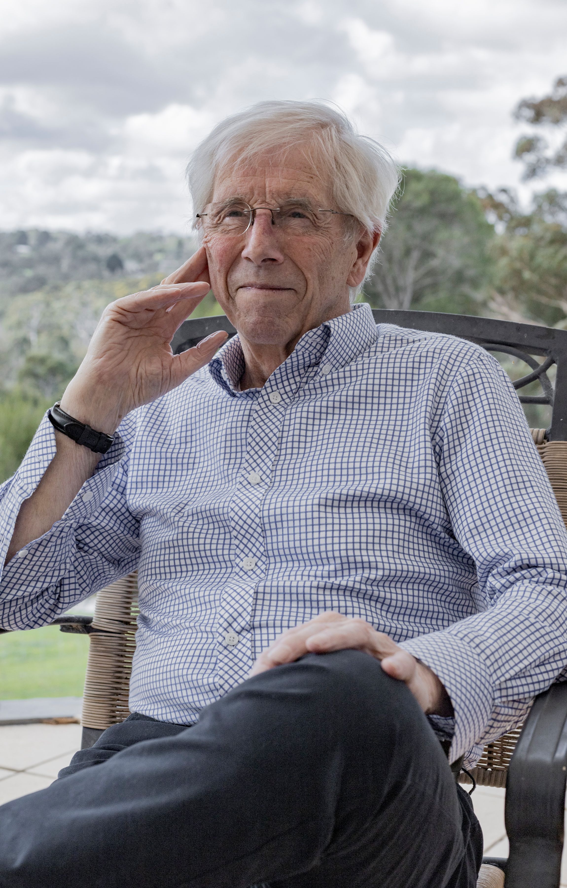 Elderly man in glasses and checkered shirt sitting outdoors, looking relaxed.
