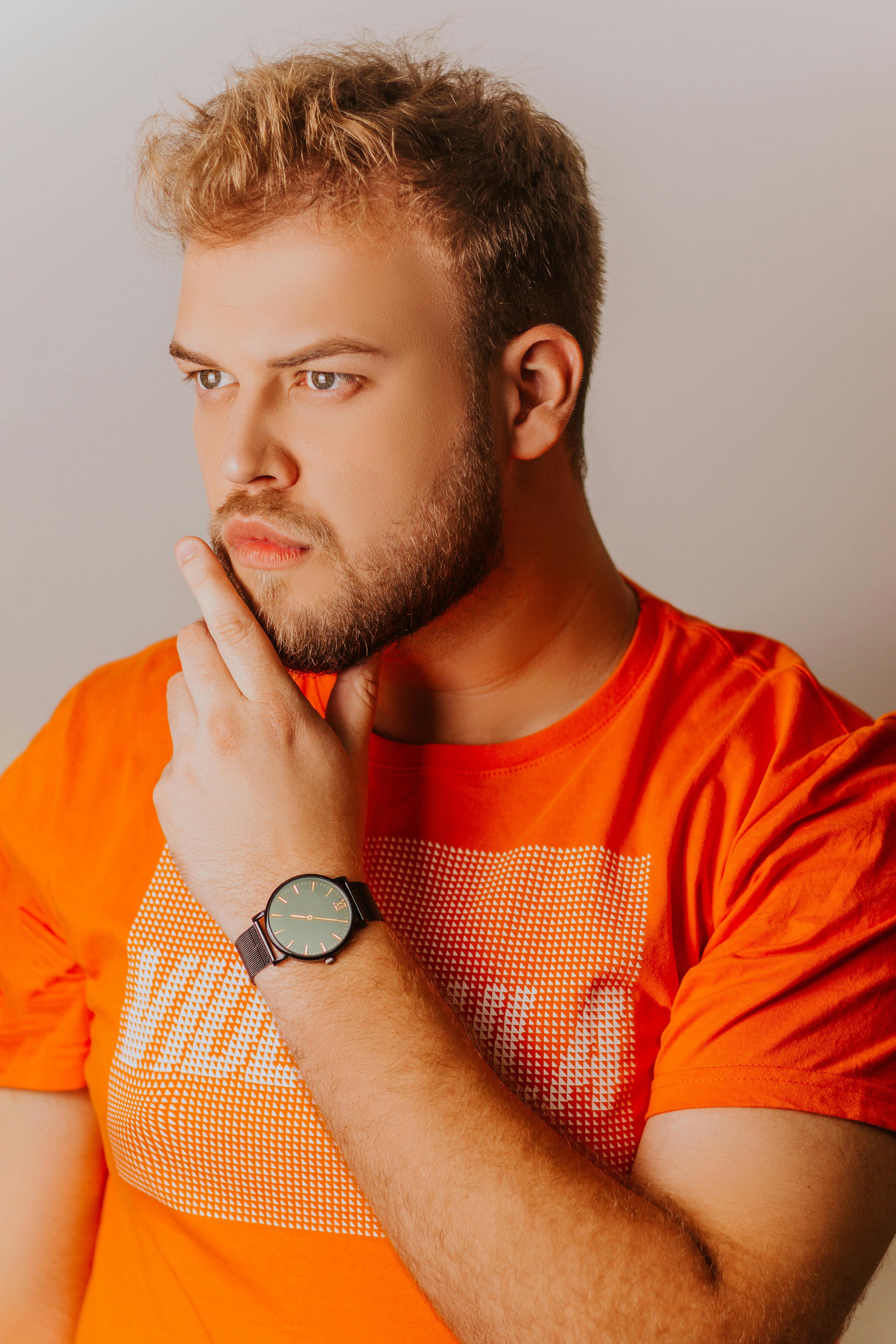 Thoughtful man with a beard wearing an orange shirt and wristwatch indoors.