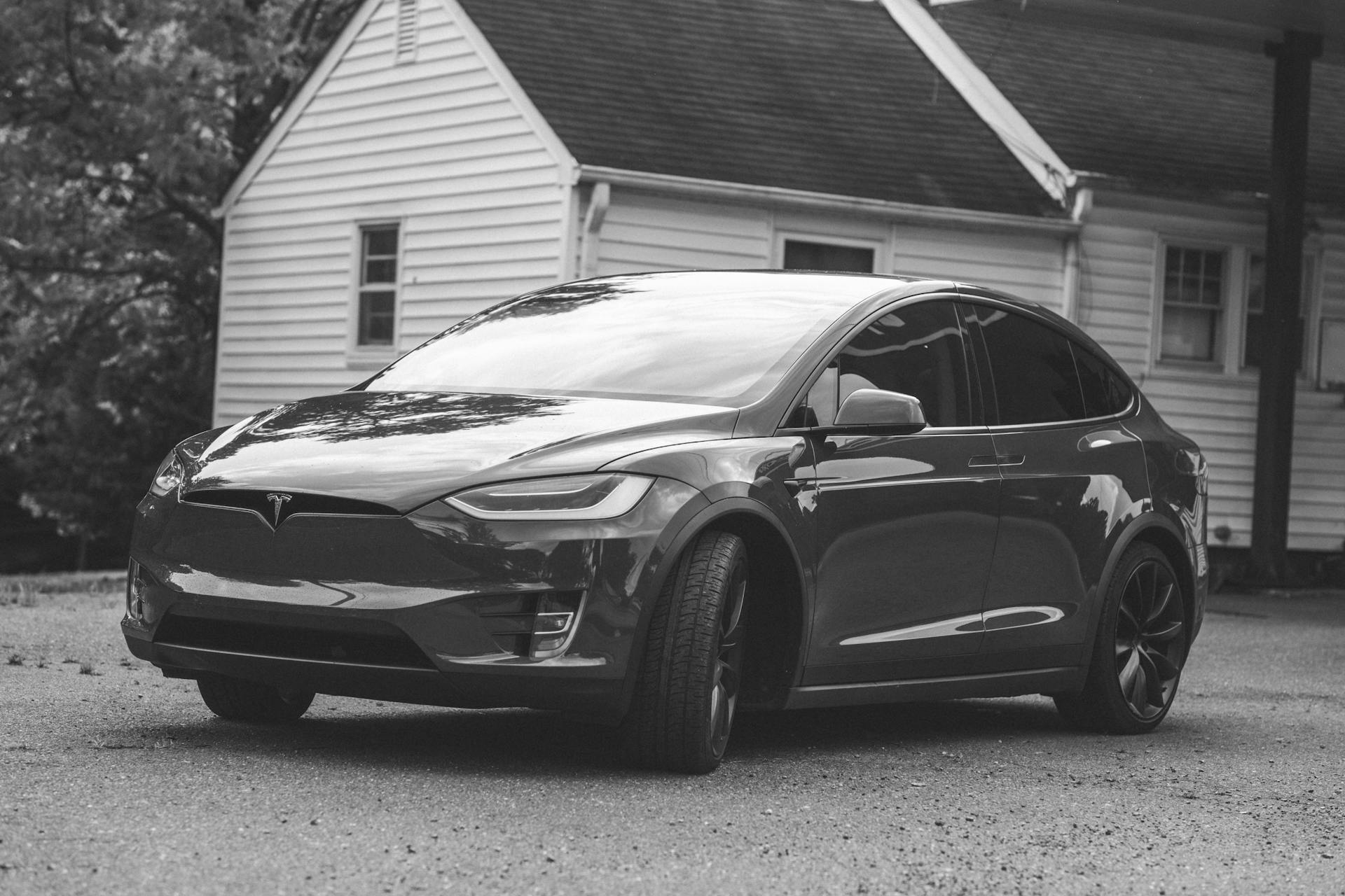 Black and white image of a Tesla car parked in front of a wooden house in a tranquil setting.