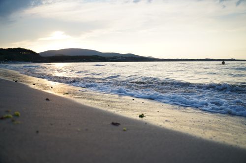 Free Tranquil view of waves crashing on a sandy beach with a stunning sunset backdrop and distant mountains. Stock Photo