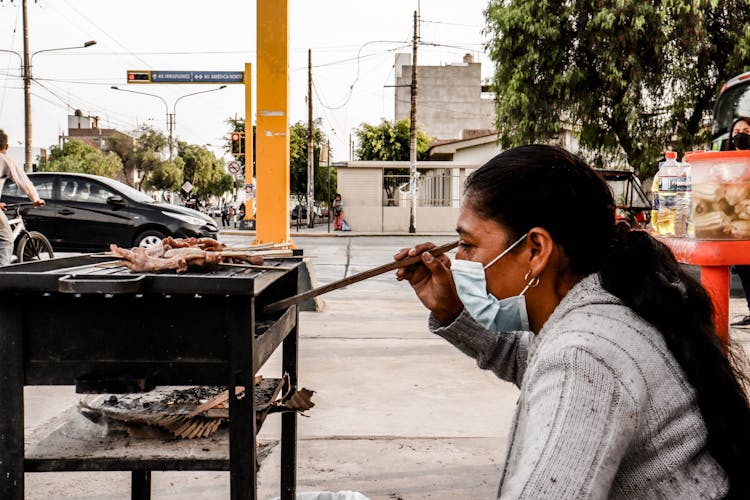 A Woman Wearing Face Mask Grilling Meat On The Side Of The Street