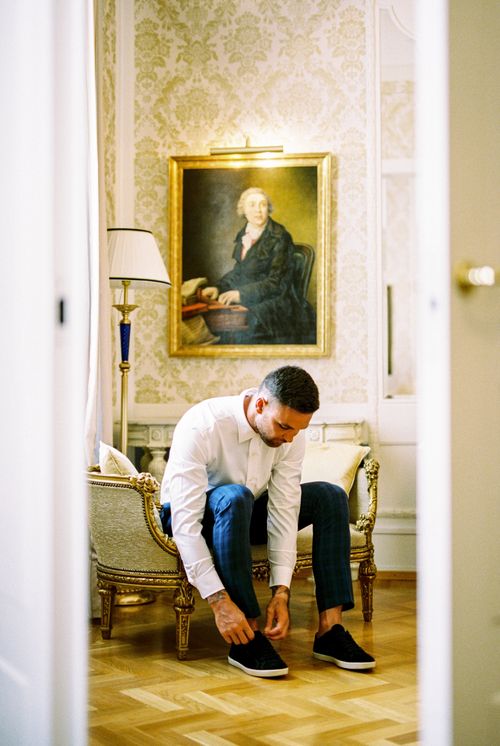 Free A stylish man sitting in an ornate room tying his shoes beneath a classic painting. Stock Photo