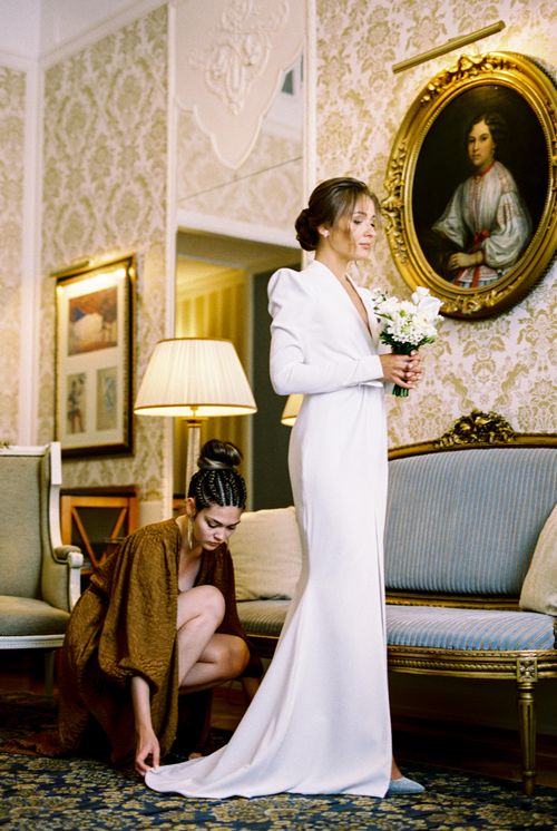Free A bride in a wedding dress with a bouquet, being attended by a woman in a lavish room. Stock Photo