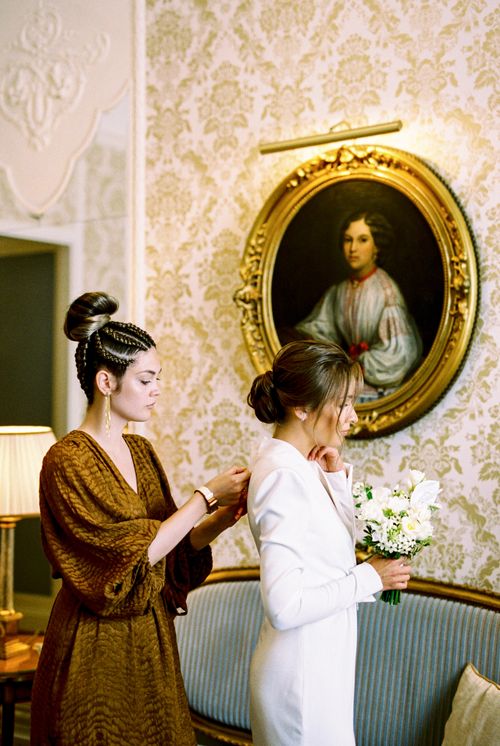 Free Two women in elegant attire prepare for a wedding, with a vintage portrait in the background. Stock Photo