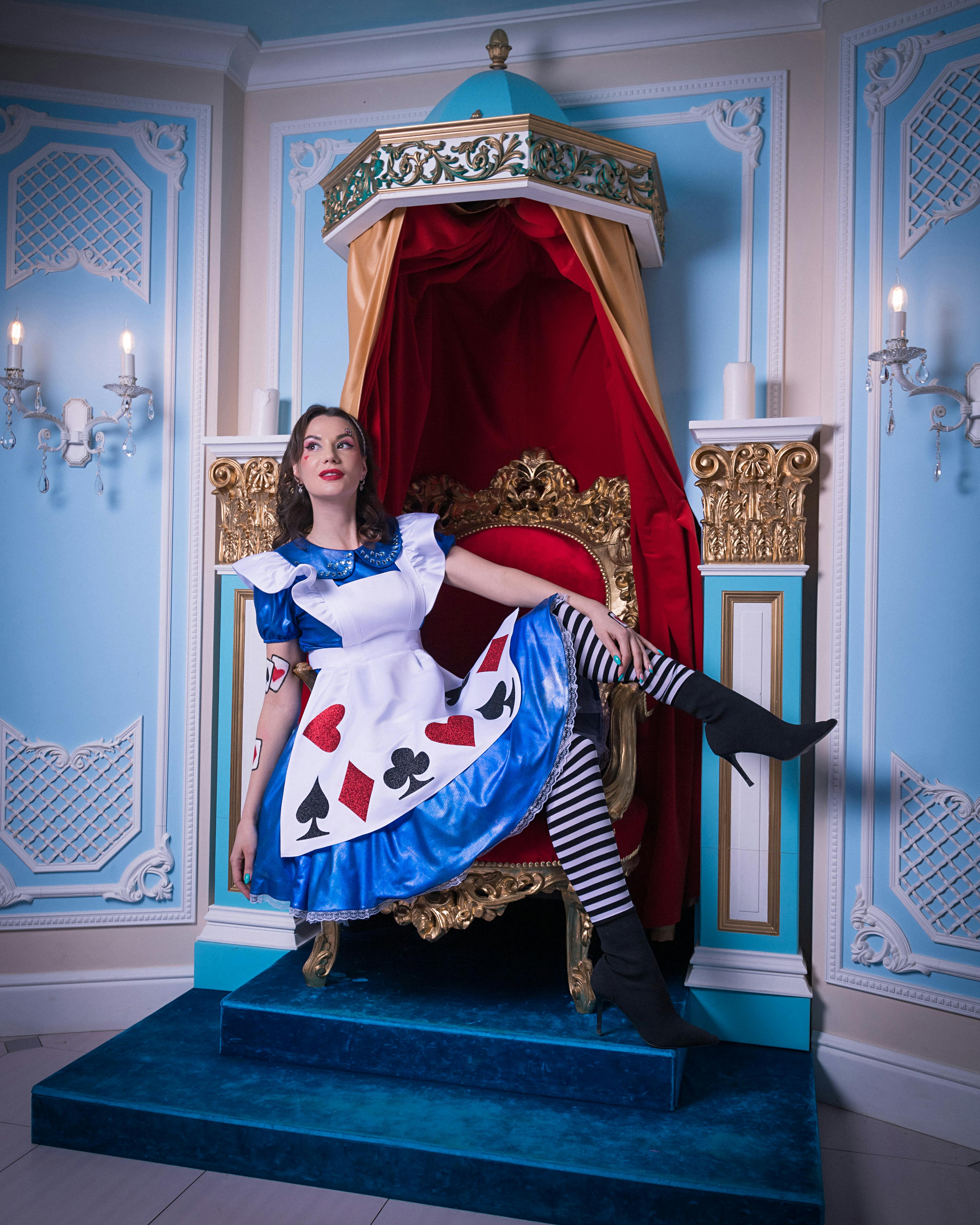 A woman in a whimsical card-themed costume poses elegantly on a lavish throne indoors.