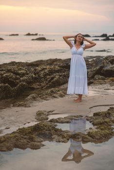 A woman in a white dress poses on a rocky beach at dusk, reflecting tranquility and relaxation.