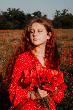 A woman in a red dress holds a bouquet of poppies, standing in a sunlit field with eyes closed.