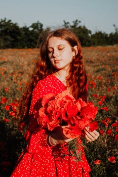 A young woman in a red dress stands serenely holding a bouquet of poppies in a sunlit meadow.