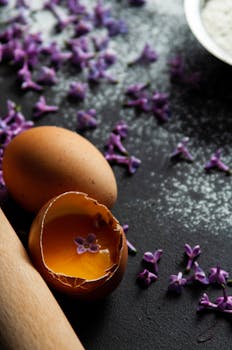 Close-up of eggshell with yolk and lilac flowers on a flour-dusted surface.