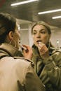 A Woman in Brown Jacket Applying a Lipstick while Looking at the Mirror
