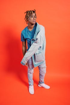 Stylish person with dreadlocks in denim outfit posing indoors with orange backdrop.