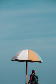 Backpacker stands under a striped sunshade against a clear blue sky in Puducherry, India.
