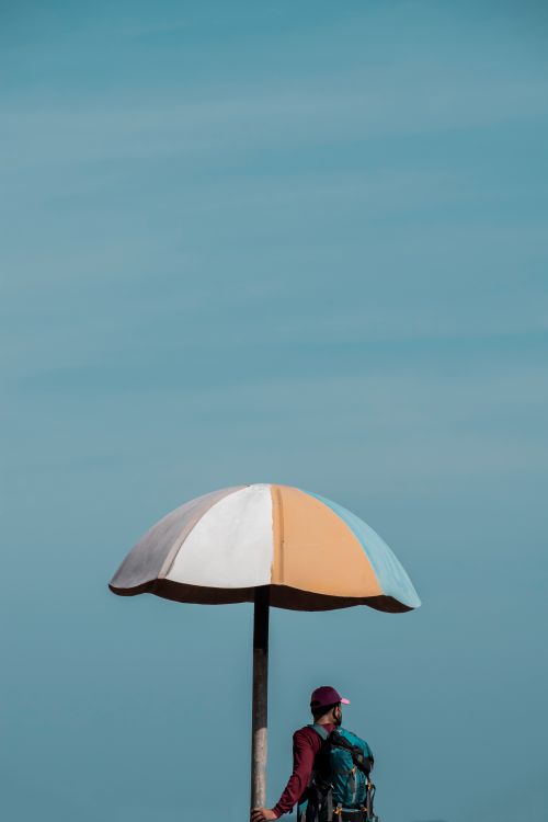 Free Backpacker stands under a striped sunshade against a clear blue sky in Puducherry, India. Stock Photo