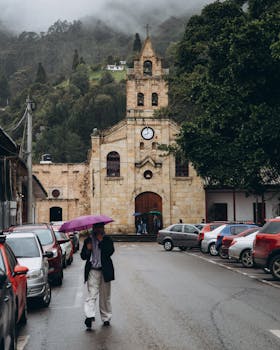 A person walks with an umbrella near a historic church with misty mountains in the background.