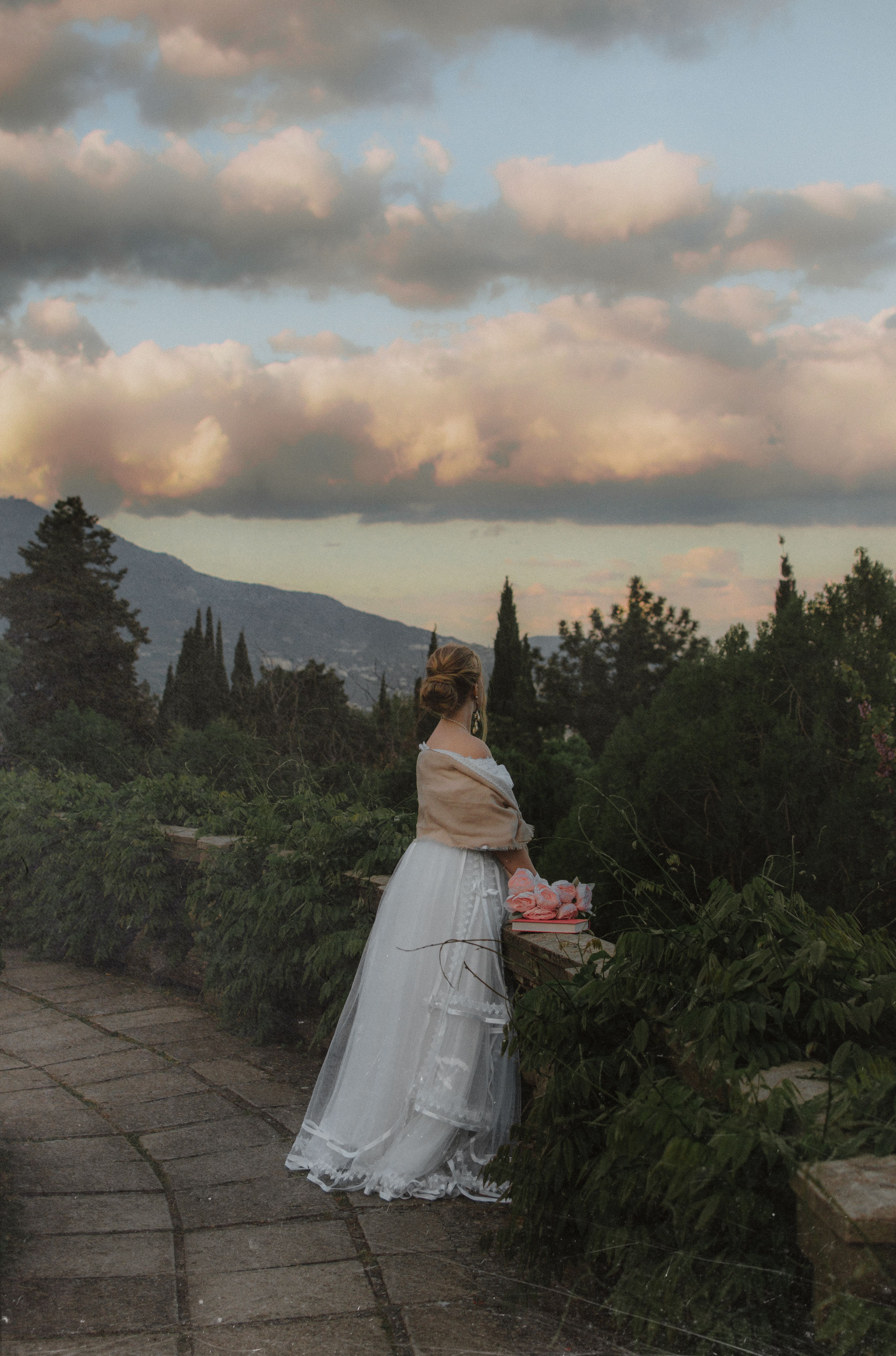A woman in a white dress stands by a garden railing, surrounded by nature at sunset.