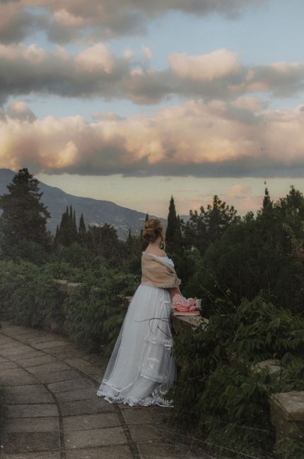 A woman in a white dress stands by a garden railing, surrounded by nature at sunset.