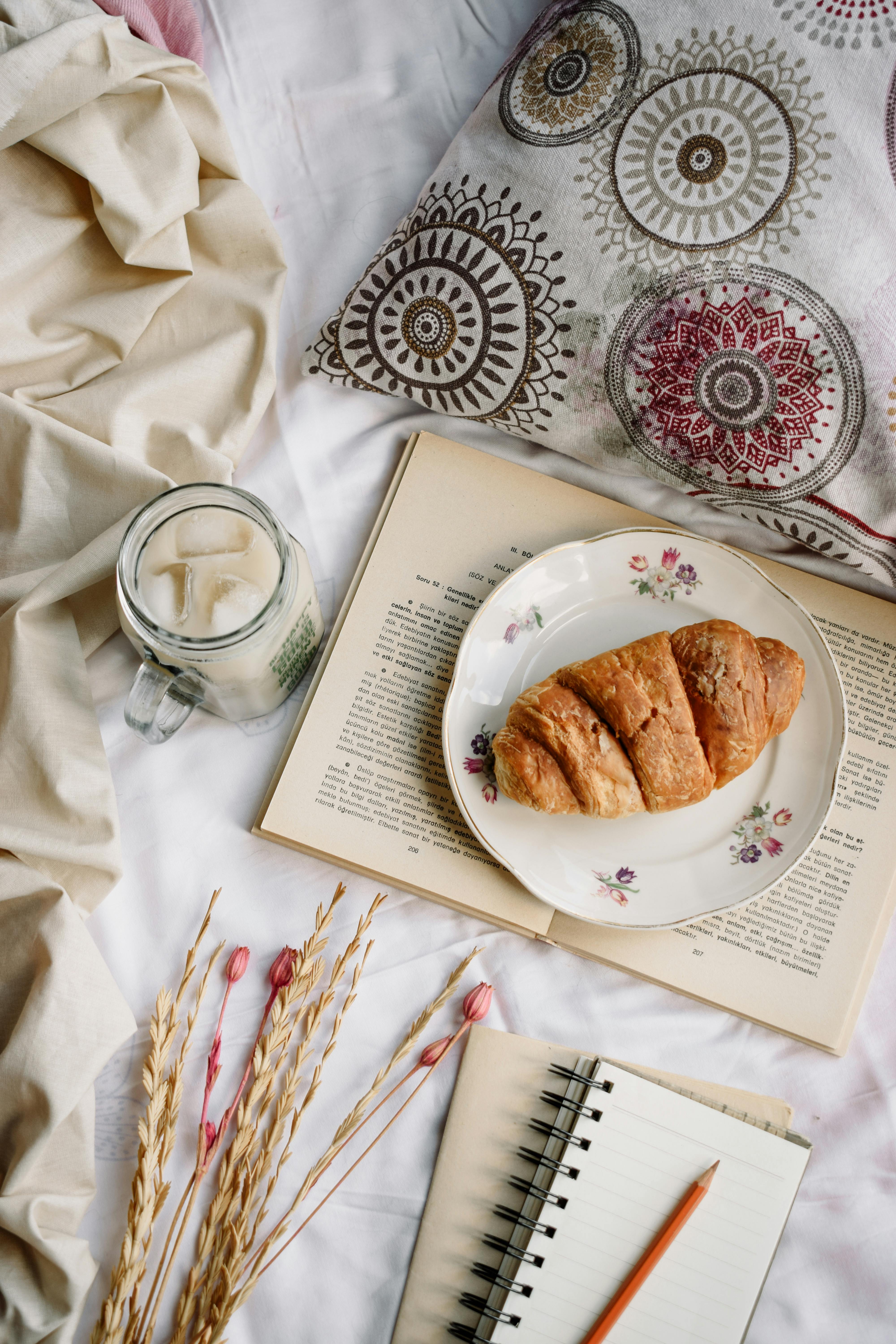 croissant on saucer on top of a book beside a cold drink