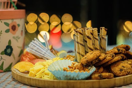 Close-up of a snack tray with cookies, chocolate sticks, and nuts, against a bokeh-lit background.
