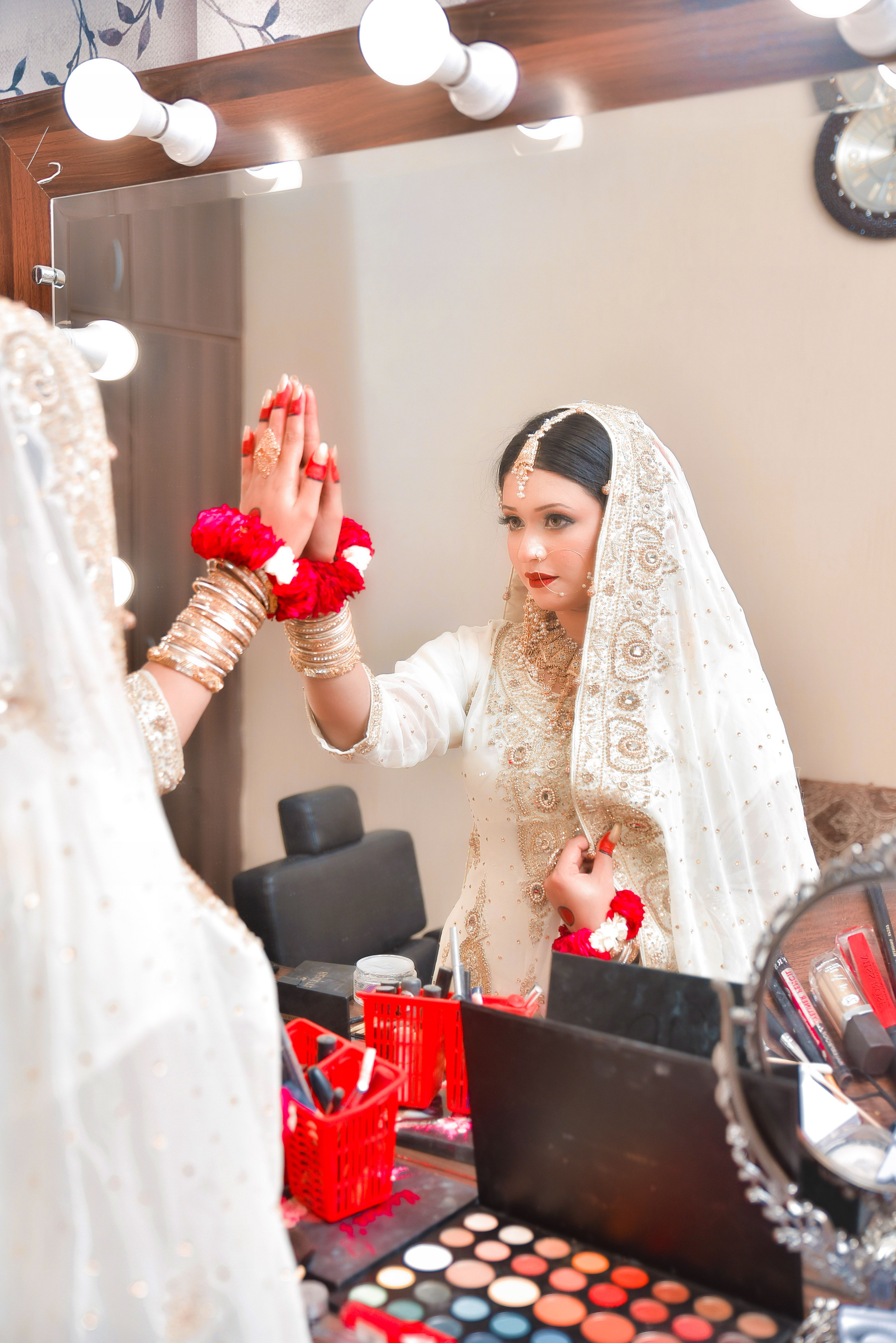 A bride in traditional attire with intricate embroidery stands before a mirror.