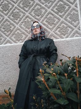 A fashionable woman in a trench coat and sunglasses poses stylishly against a textured wall.
