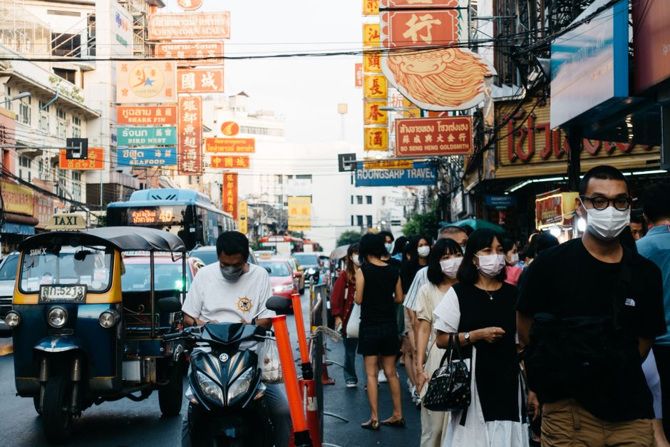 People wearing face masks walk in bustling Chinatown street, amidst traffic and vibrant signs.
