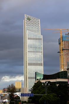 A tall modern skyscraper in a Chinese city with dramatic cloudy sky and urban structures.