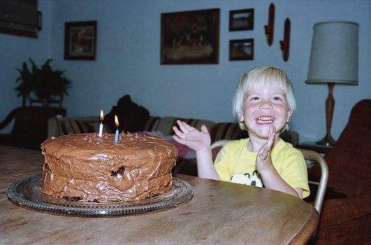 Smiling child clapping next to chocolate cake with candles indoors.