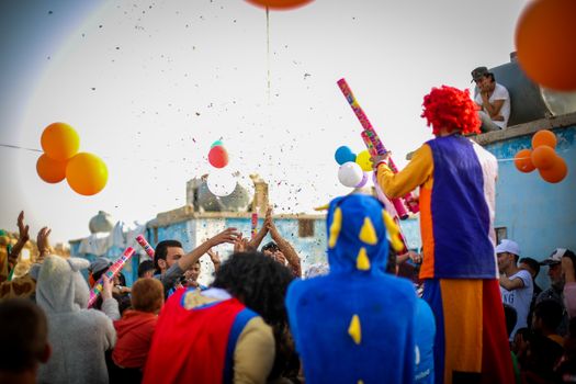 A lively street celebration in Idlib with clowns, costumes, and balloons, creating a joyful atmosphere.
