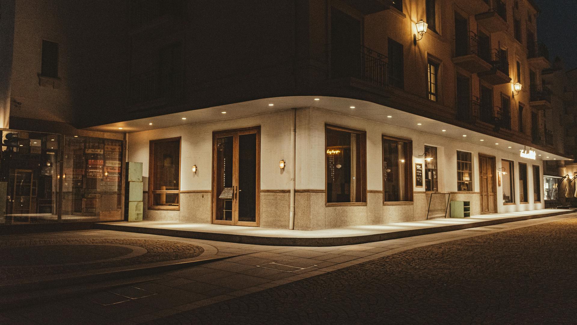 Warmly lit corner building exterior with large windows and sconces at night.