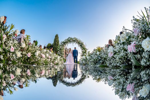 A romantic outdoor wedding ceremony featuring a floral arch, bride and groom reflection, and guests in a garden setting.