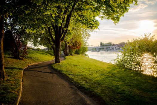 Beautiful sunset view of a riverside park with trees, a path, and a bridge in Compiègne, France.