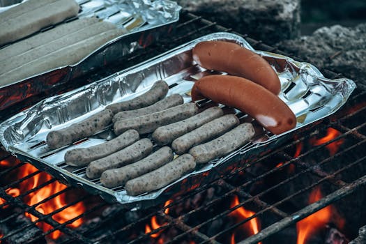 Close-up of assorted bratwurst and sausages cooking over an open fire on aluminum trays.
