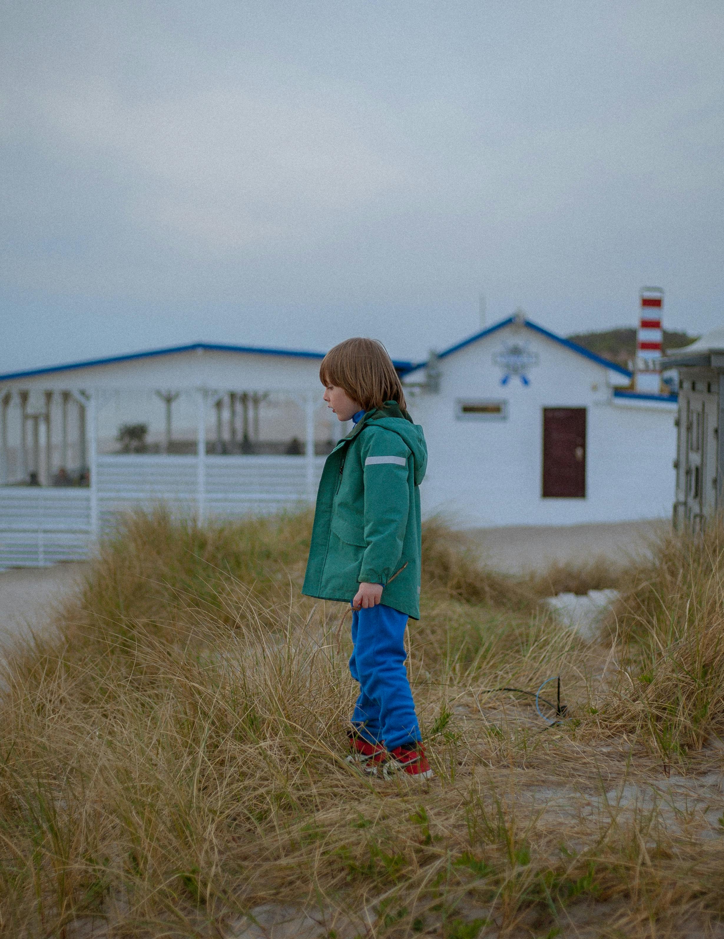 A young boy in a green jacket stands amidst sand dunes with a seaside backdrop.