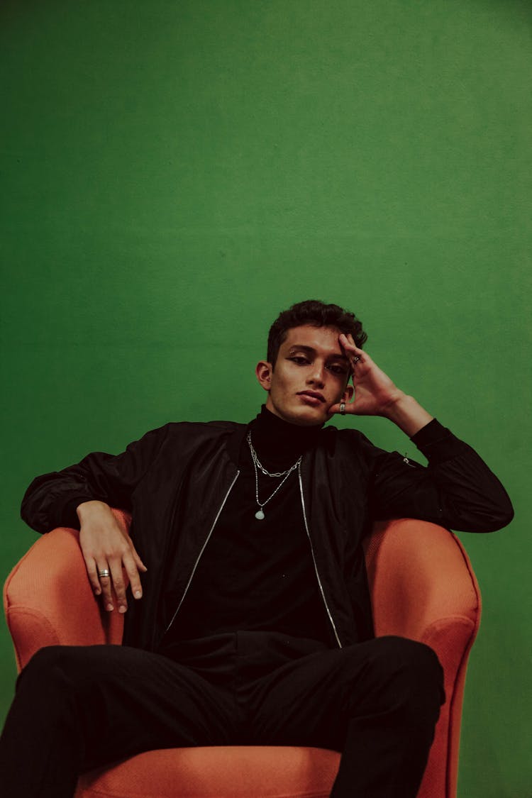 Young Man In Black Outfit With Necklace Sitting In Armchair