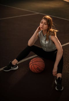 A young woman in athletic attire sits on a basketball court with a basketball, deep in thought.