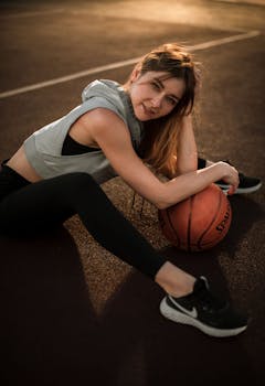 Casual young woman in activewear sitting on outdoor court with basketball during daytime.