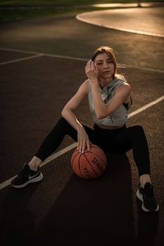 A young woman in sportswear sitting on a basketball court with a ball during sunset.