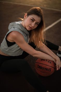 A young woman sitting on an outdoor basketball court, holding a basketball under warm sunlight.
