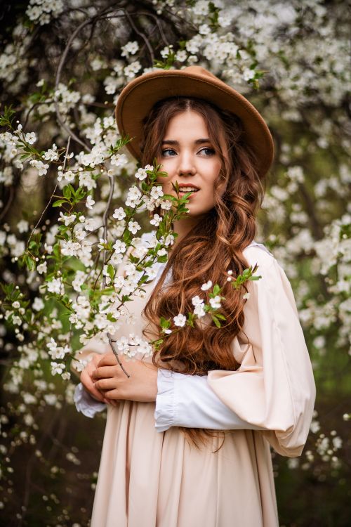 Free A young woman in a flowy dress and hat stands among blossoming trees, surrounded by white flowers. Stock Photo