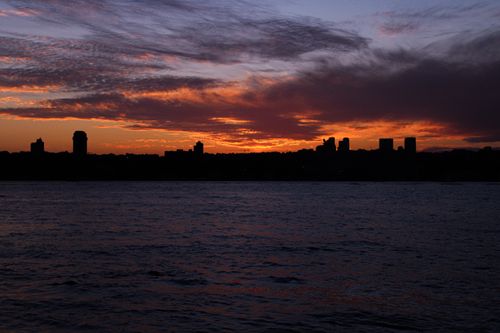 Free Silhouetted skyline and vivid sunset over calm waters create a dramatic urban landscape. Stock Photo