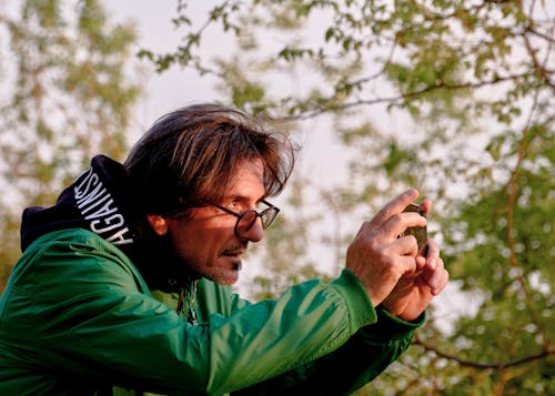 Man in Green Jacket Wearing Eyeglasses Holding a Smartphone 