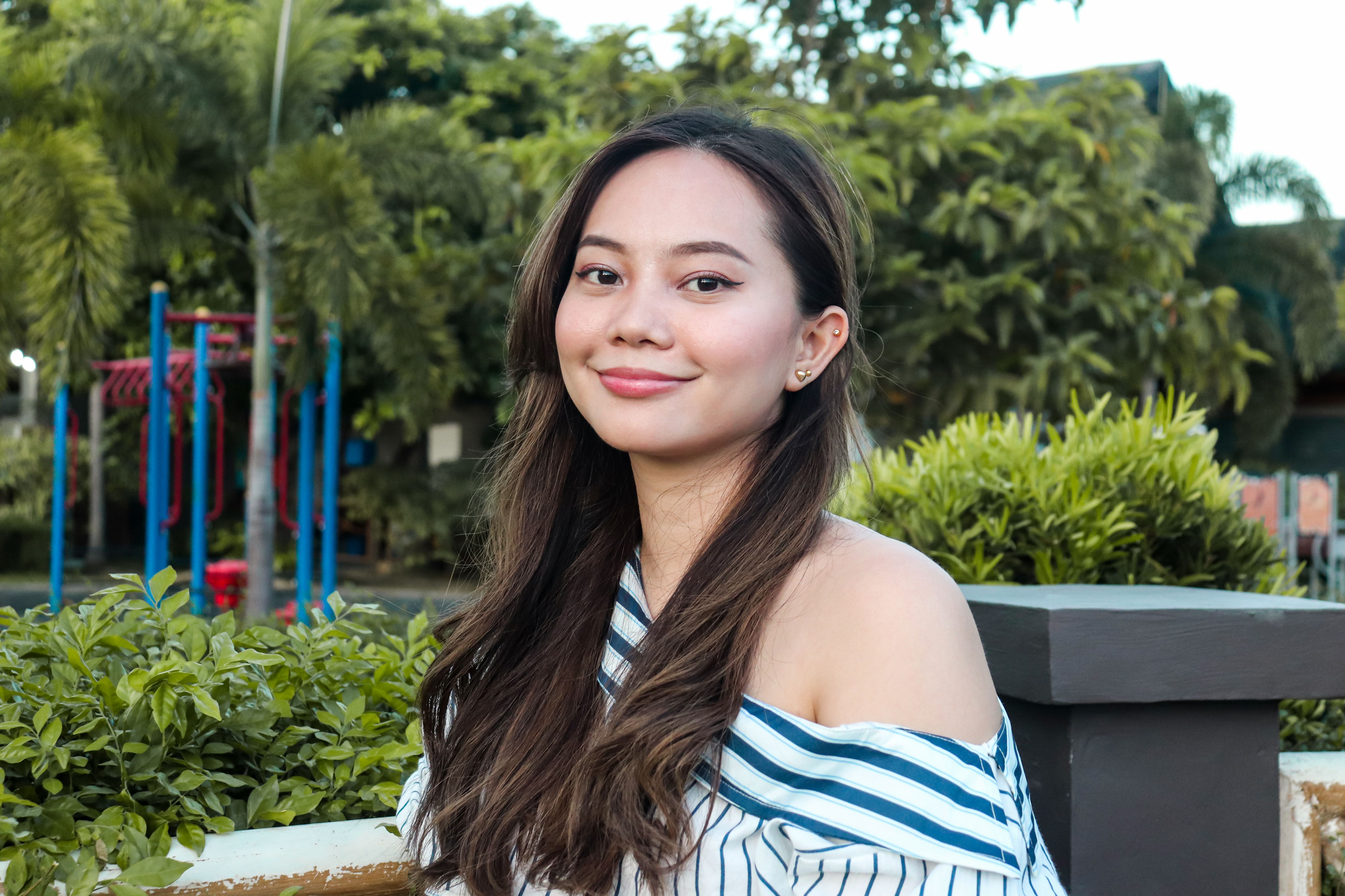 A smiling woman with long hair in an off-shoulder top, outdoors in a park setting.