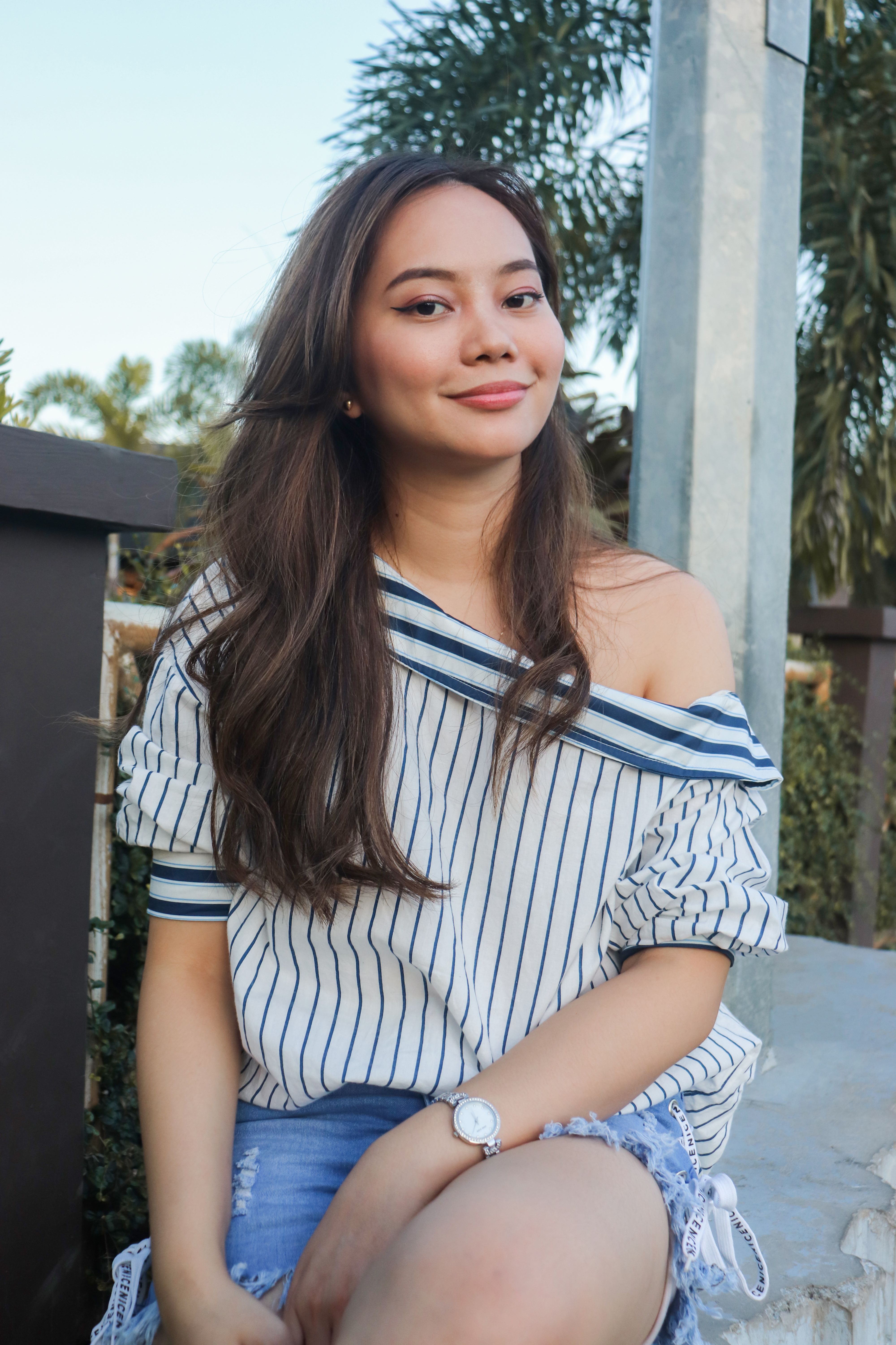 Young woman wearing a striped shirt and wristwatch, sitting outdoors with a cheerful smile.