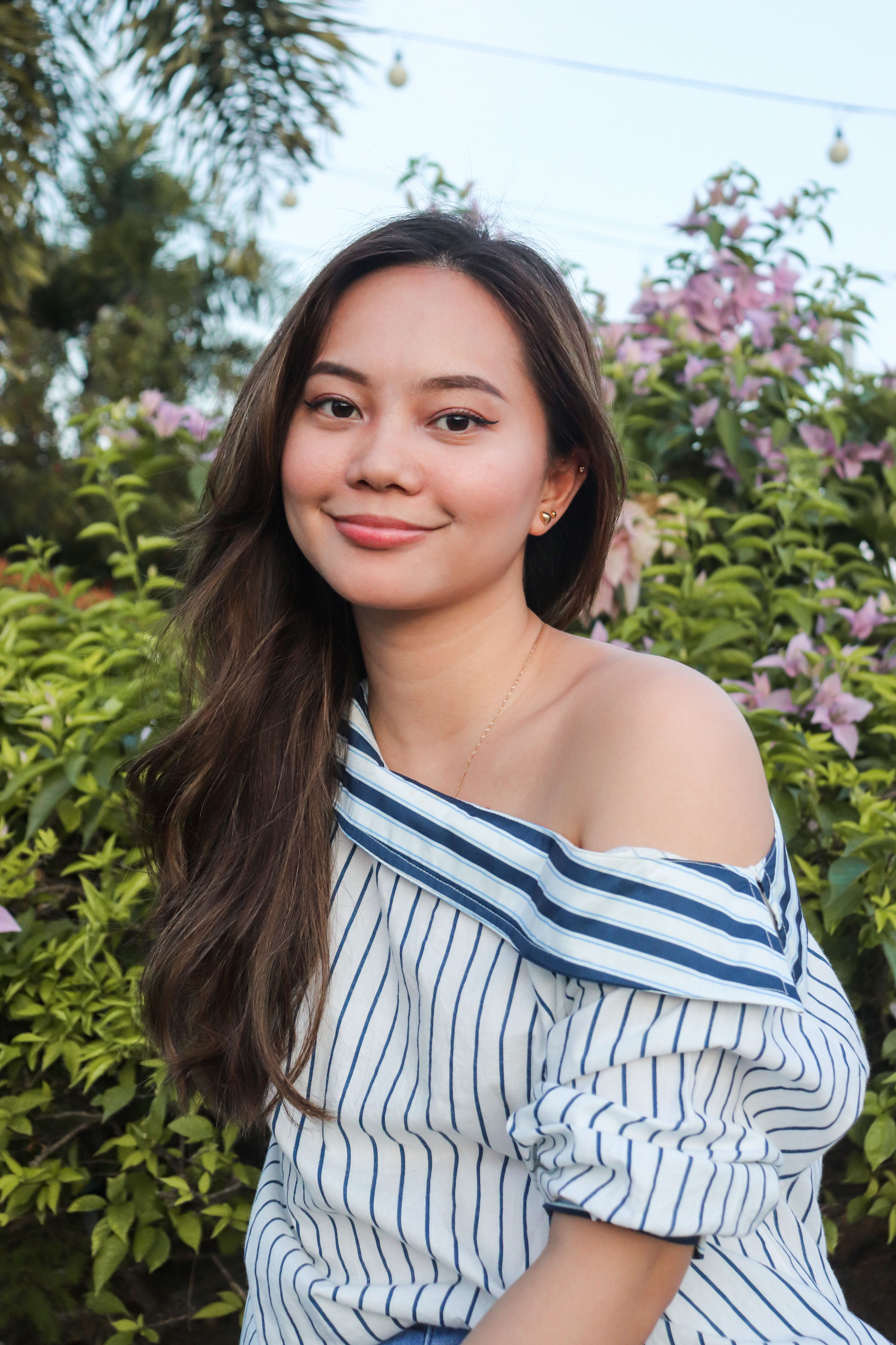 A young woman in a striped blouse smiling outdoors in a vibrant garden setting.