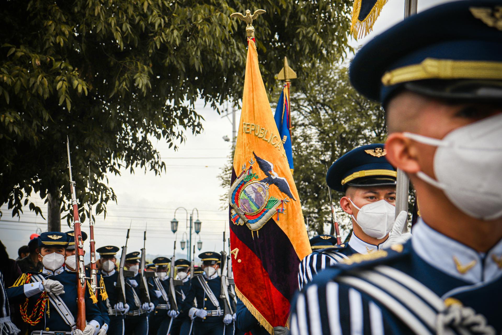 Military personnel in uniforms with masks march in an Ecuadorian parade featuring the national flag.