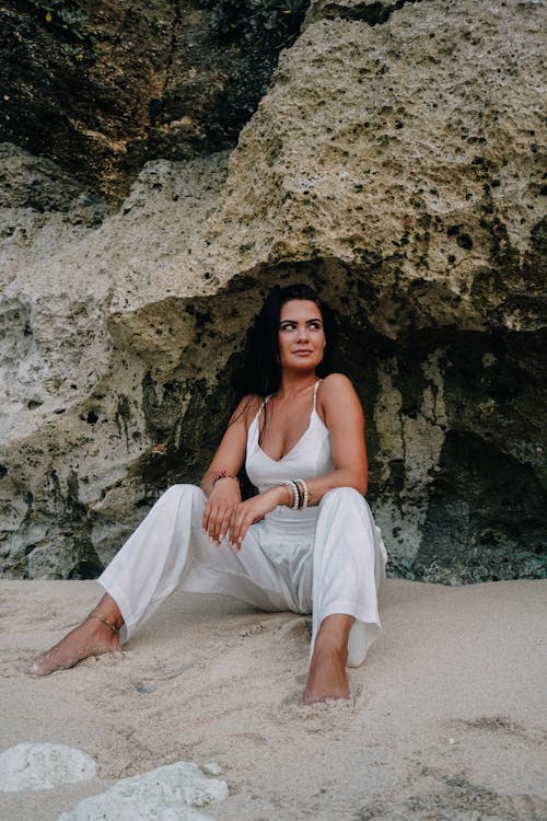 A Woman in White Spaghetti Strap Jumpsuit Sitting on Sand Near Rock Formation